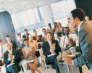 Businessman Standing at a Podium and Giving a Speech to a Conference Room Full of Delegates